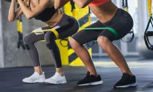 Dwie kobiety wykonują ćwiczenia z gumami oporowymi na udach, przypominające przysiady. Nie jest to hack squat maszyna, ale ćwiczenie angażujące podobne partie mięśni.