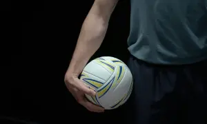 Dłoń zawodnika trzyma piłkę do siatkówki. Ciemne tło podkreśla biało-żółto-niebieską piłkę, gotową do gry w siatkówkę mężczyzn plusliga.