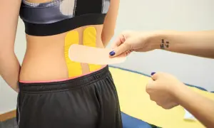 Naklejanie tejpy na plecy w celu wsparcia mięśni. Dłoń z tatuażem "You can do it" przygotowuje plaster.
