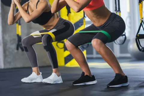 Dwie kobiety wykonują ćwiczenia z gumami oporowymi na udach, przypominające przysiady. Nie jest to hack squat maszyna, ale ćwiczenie angażujące podobne partie mięśni.