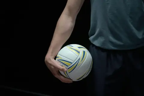 Dłoń zawodnika trzyma piłkę do siatkówki. Ciemne tło podkreśla biało-żółto-niebieską piłkę, gotową do gry w siatkówkę mężczyzn plusliga.