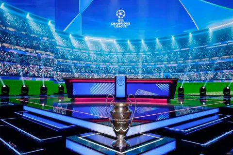 Trofeum Ligi Mistrzów na scenie z widownią w tle. Tuż obok ekran z napisem "Liga Mistrzów", który może wyświetlać tabelę wyników.