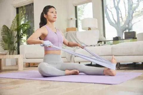 Kobieta ćwiczy pilates w domu, siedząc na macie i napinając taśmę oporową.