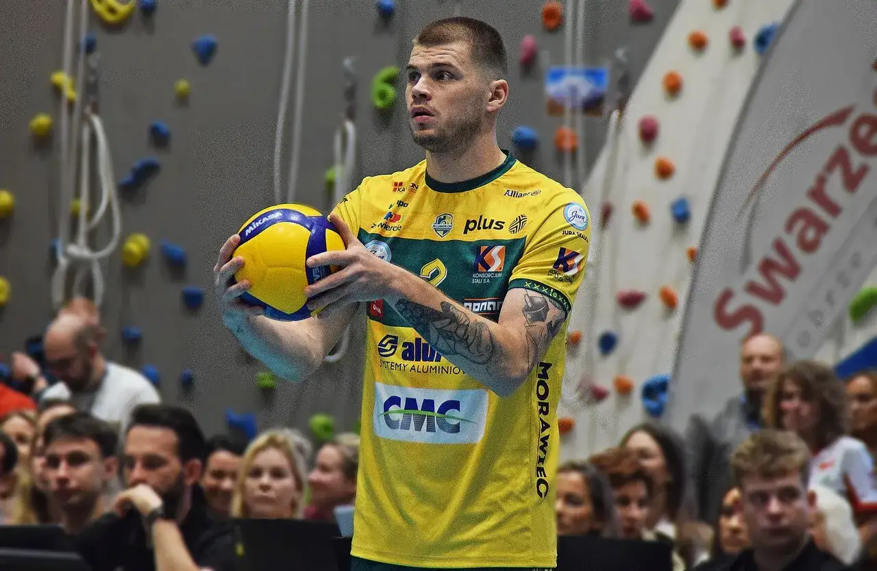 Bartek Kwolek, siatkarz w żółtej koszulce, trzyma piłkę do siatkówki. W tle ściana wspinaczkowa i publiczność.